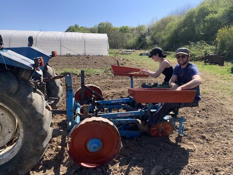 Plantation de pommes de terre Deux personnes souriantes travaillent ensemble sur un tracteur pour planter des pommes de terre dans un champ ensoleillé, entourées de verdure et d’un ciel bleu radieux. Une belle journée de collaboration et de partage au cœur de la nature !